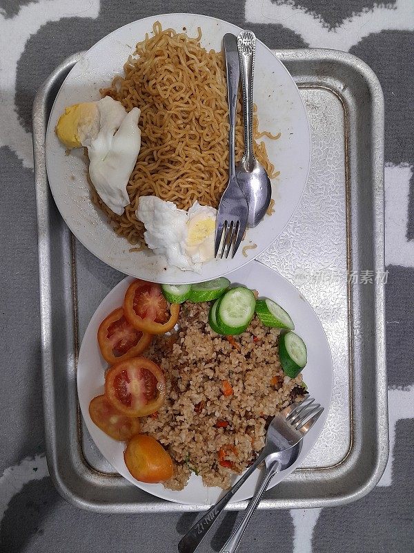 a plate of fried rice and a plate of instant noodles Fried Indomie is a very popular food in Indonesia which is served with fried eggs, sliced ​​cucumbers and tomatoes to add to the taste of the food.